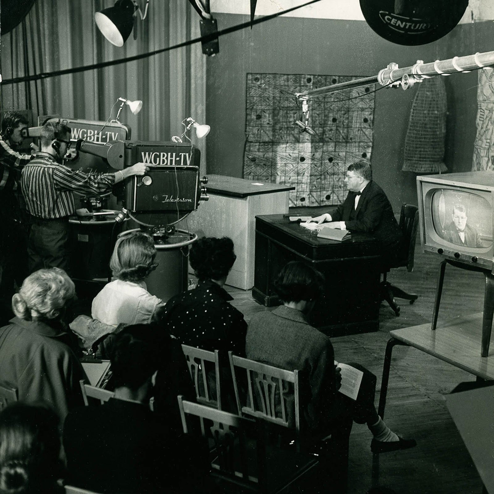 A view of a WGBH broadcast. A man sits behind a desk while being filmed by cameras that read 